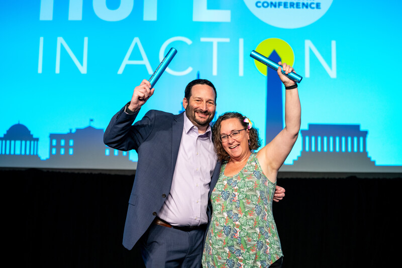 man in a sports coat on the left and a woman in a floral shirt on the right, both lifting batons in celebration